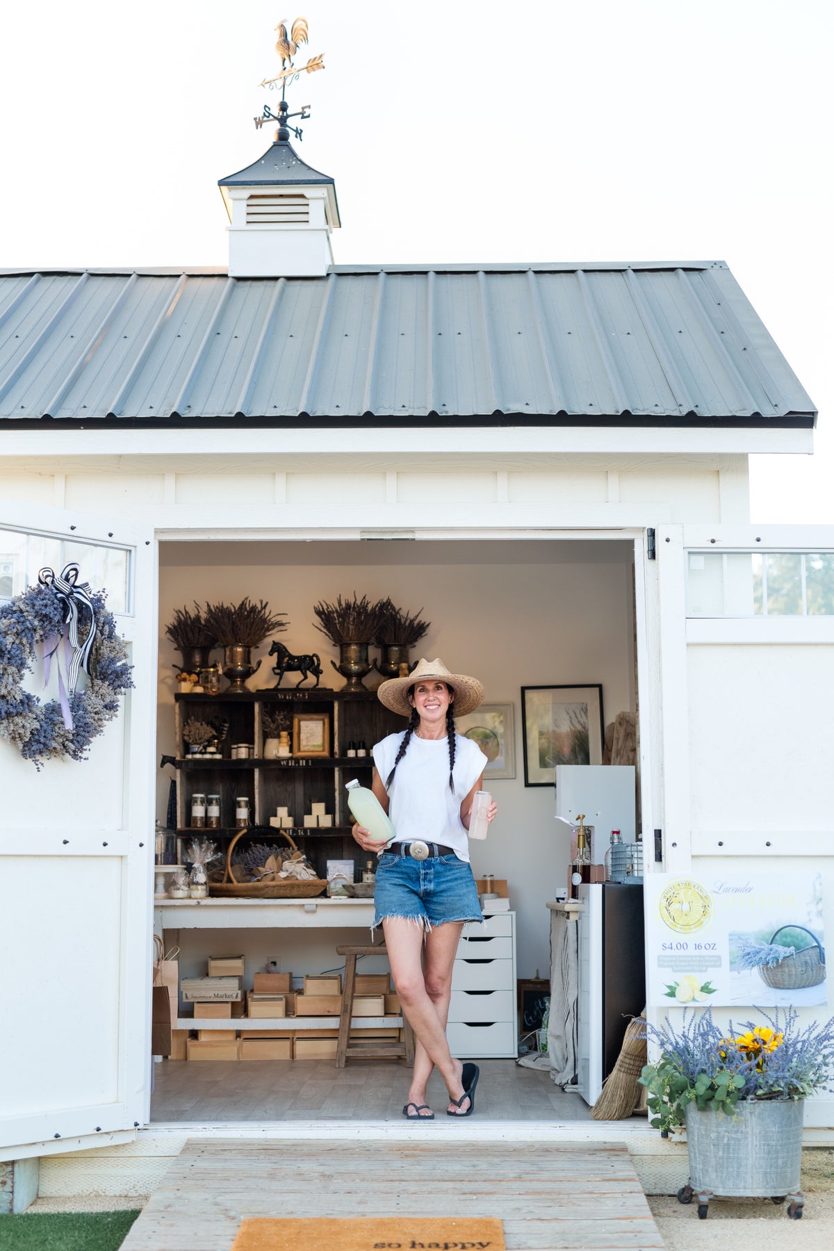 A woman wearing a straw hat, white t-shirt, and denim shorts stands in the doorway of a small white rustic shop with a gray metal roof, holding a green bag. The shop interior is visible behind her, decorated with shelves, plants, and baskets.